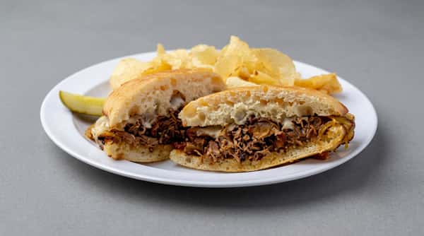 Grilled steak and cheese sub sandwich with potato chips and a pickle spear on a white plate.