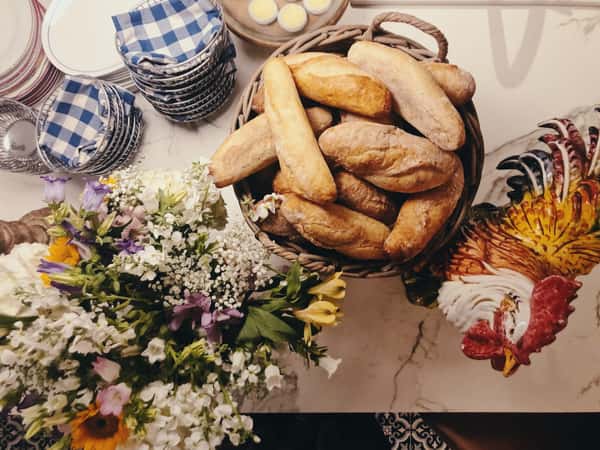 Basket of fresh baguettes beside a bouquet of colorful flowers and a decorative ceramic rooster on a marble countertop.