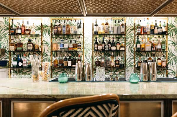Elegant bar with tropical leaf-patterned wallpaper, shelves of liquor bottles, and a marble countertop with cocktail-making tools.