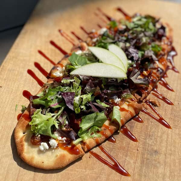 Flatbread topped with mixed greens, goat cheese, bacon, sliced green apple, and drizzled with a balsamic or barbecue glaze — a fresh and savory gourmet dish.