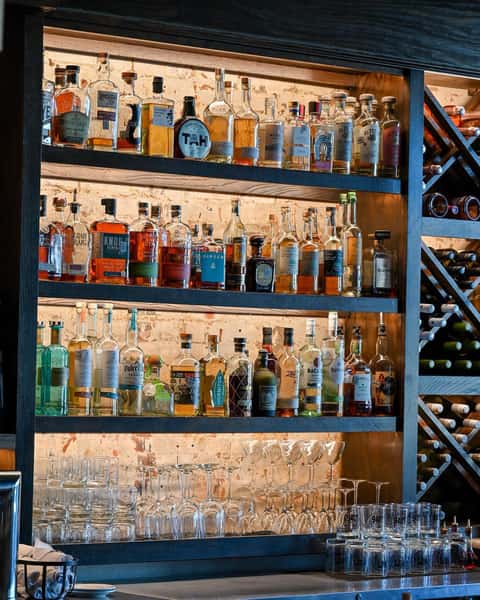 Backlit bar shelf stocked with assorted liquor bottles and glassware, adjacent to a vertical wine rack filled with bottles.