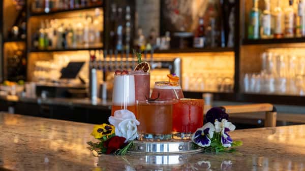 Colorful cocktail flight featuring five different drinks, garnished with fruit and herbs, arranged on a bar counter with flowers.