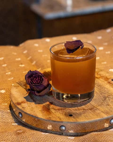 Brown cocktail served on the rocks in a clear glass, garnished with a dried purple rose petal, next to a dried rose on a wood tray.
