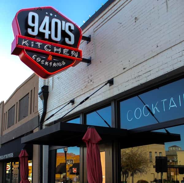 Exterior view of 940's Kitchen & Cocktails featuring a bold red and black neon sign above large front windows.