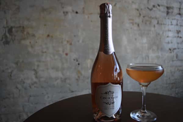Bottle of Le Grand Courtâge Brut Rosé with a filled coupe glass on a round table against a rustic brick wall background.