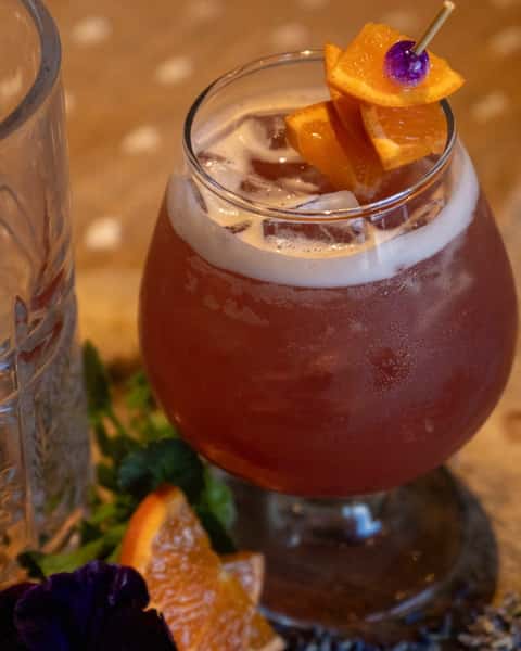 Fruity cocktail served over ice in a stemmed glass, garnished with orange slices and a purple cherry on a skewer.