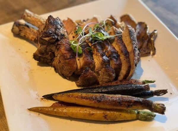 Grilled pork chop sliced and served with roasted rainbow carrots on a square white plate, garnished with microgreens.