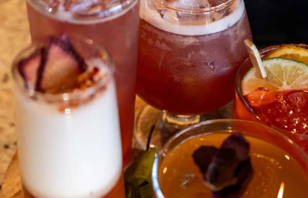 Assorted colorful cocktails garnished with citrus slices, berries, and foam served in a mix of glasses on a wooden board.