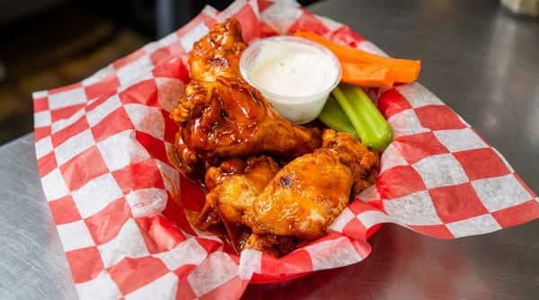Chicken wings in a checkered paper cup with carrot sticks, celery, ranch dressing, and a blurry glass of beer.