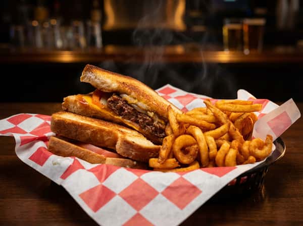 Patty melt with melted cheese, grilled onions, and a side of curly fries in a red and white checkered basket.