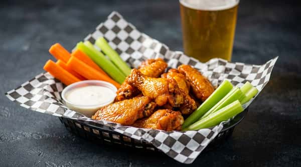 Chicken wings with celery sticks, baby carrots, and two cups of ranch dressing, served in a red and white checkered paper.