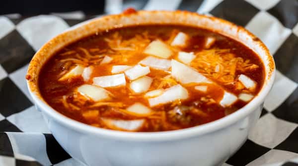 Bowl of chili with shredded cheese and diced white onions, on a black and white checkered paper liner.