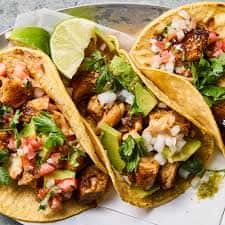 Street-style tacos with grilled meat, avocado, onions, and cilantro in soft corn tortillas, served with lime wedges for an authentic Mexican restaurant presentation.