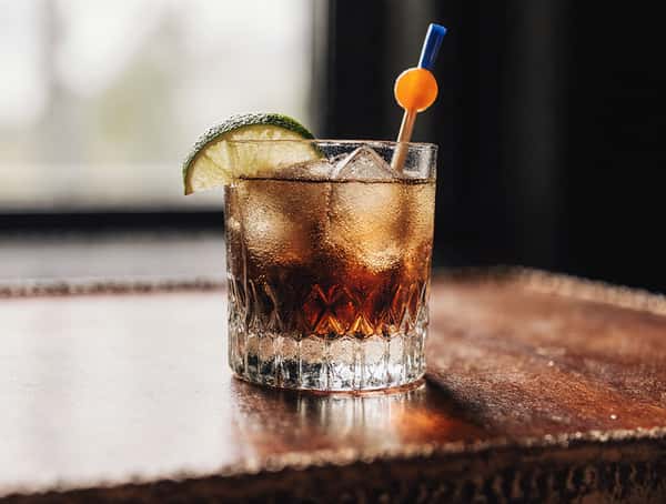 Craft cocktail with lime wedge and ice served in a rocks glass on a wooden bar table.