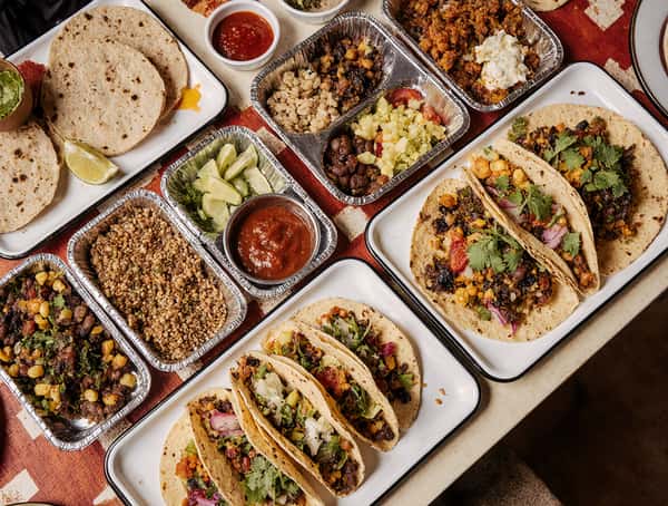 Colorful spread of Mexican street tacos on corn tortillas topped with beans, corn, cilantro, and diced vegetables, served alongside trays of grains, black beans, salsa, lime wedges, and fresh tortillas on a rustic restaurant table.