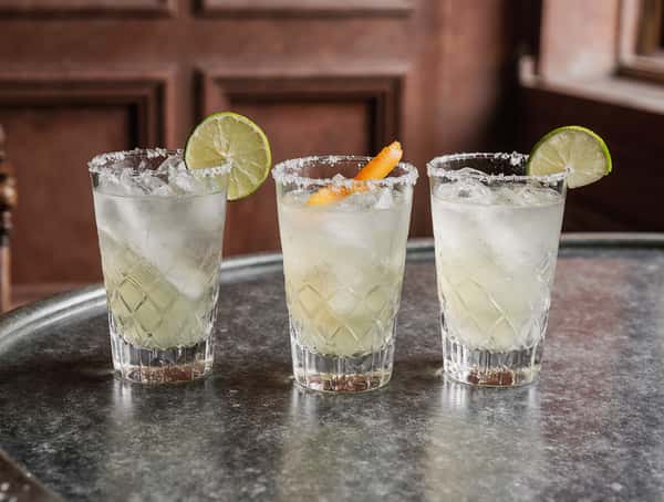 Three salt-rimmed margarita cocktails with lime and orange garnish served on a bar tray in a Mexican restaurant.
