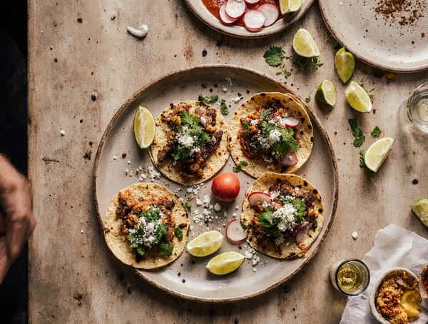 Four street tacos with seasoned meat, cilantro, radish slices, queso fresco, and lime wedges on a rustic table.