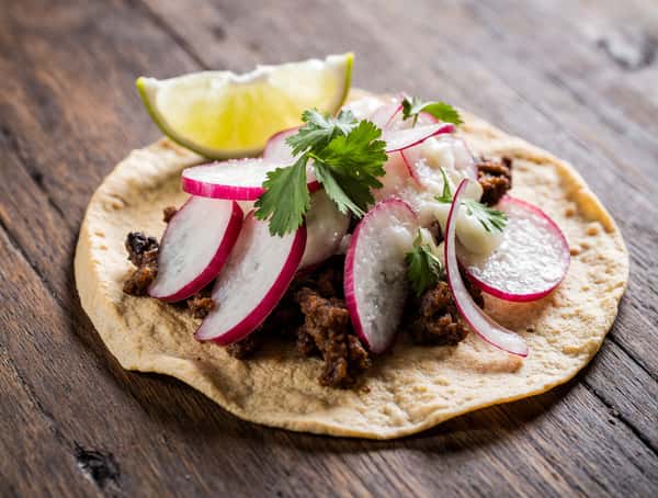 Close-up of a street-style taco with grilled meat, radishes, cilantro, crema, and lime on a corn tortilla.