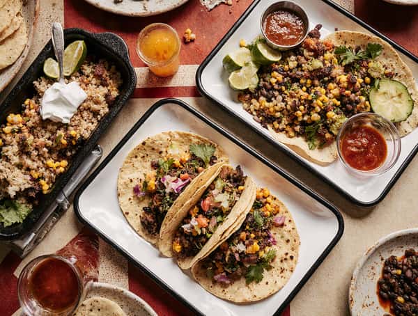 Mexican street tacos on corn tortillas topped with grilled meat, roasted corn, diced onions, and cilantro, served with fresh salsa, lime wedges, and sides of grains and beans on a colorful restaurant table.