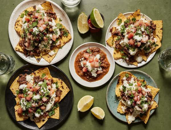 Multiple plates of loaded nachos topped with meat, melted cheese, pico de gallo, and cilantro served with salsa and lime.