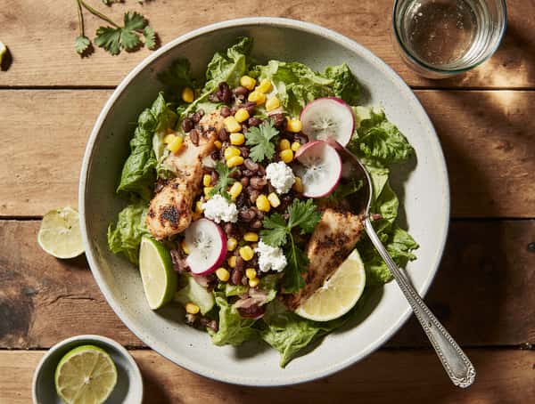 Grilled chicken Mexican salad with corn, black beans, radish slices, queso fresco, cilantro, and lime wedges.
