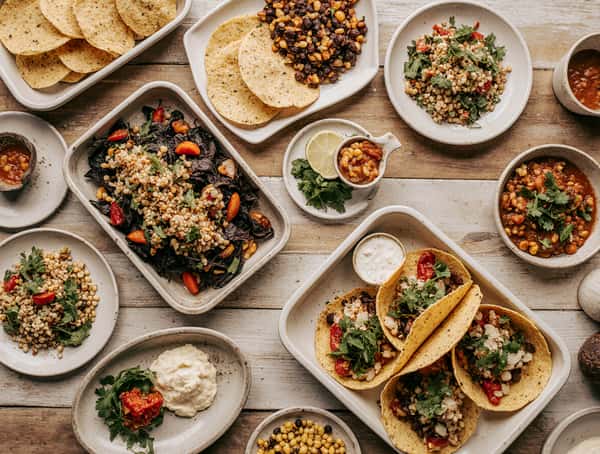 Colorful spread of Mexican dishes including tacos, tortilla chips, grain salads, roasted vegetables, and salsas on a rustic table.