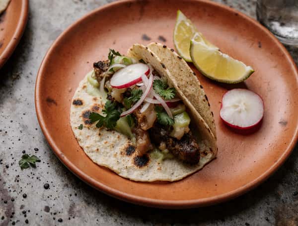 Mexican street taco on a corn tortilla topped with grilled meat, radish slices, cilantro, onions, and lime wedge.