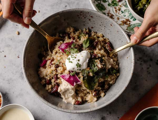 Hearty grain bowl with beans, herbs, pickled onions, greens, and creamy sauce being mixed with spoons on a marble table.