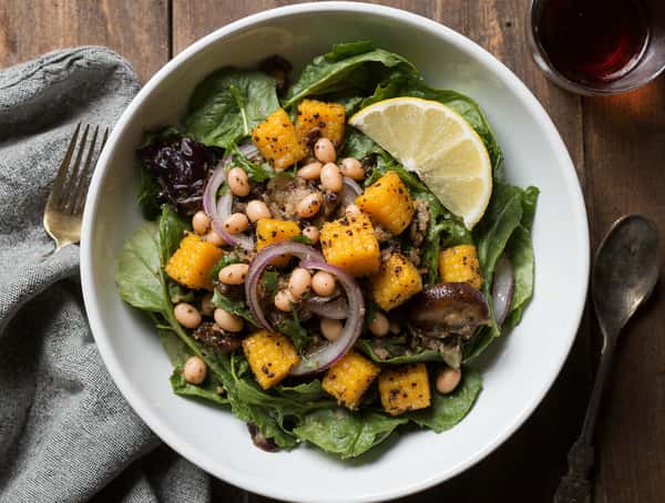 Fresh salad with leafy greens, white beans, grilled corn, red onion slices, mushrooms, and herbs, served with a lemon wedge.