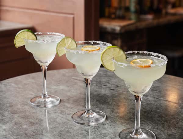 Three classic margaritas with salted rims and lime garnish in coupe glasses on a restaurant bar counter.