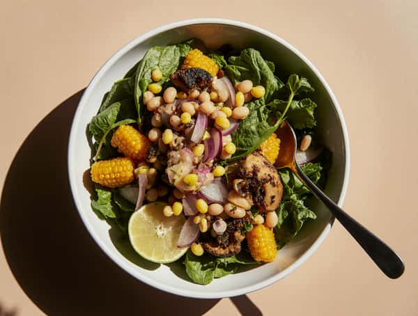 Hearty salad bowl with leafy greens, grilled mushrooms, corn, beans, red onion, and lime on a neutral background.