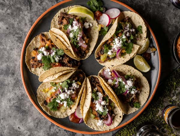 Platter of street-style tacos with grilled meat, cilantro, onion, radish slices, queso fresco, and lime wedges.