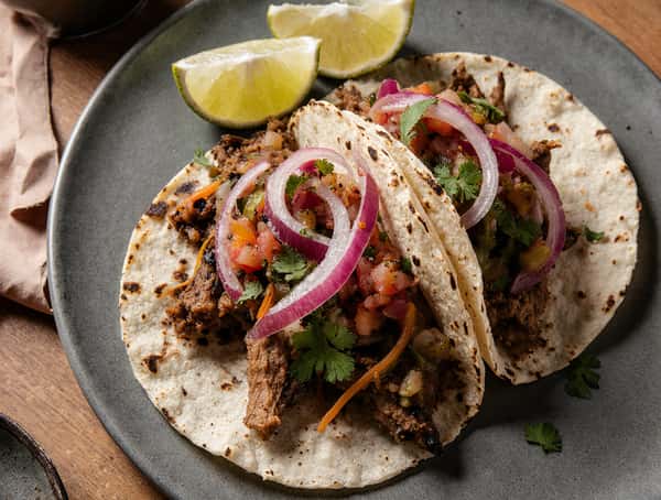 Two beef tacos with corn tortillas, red onion, cilantro, and fresh salsa served with lime wedges.