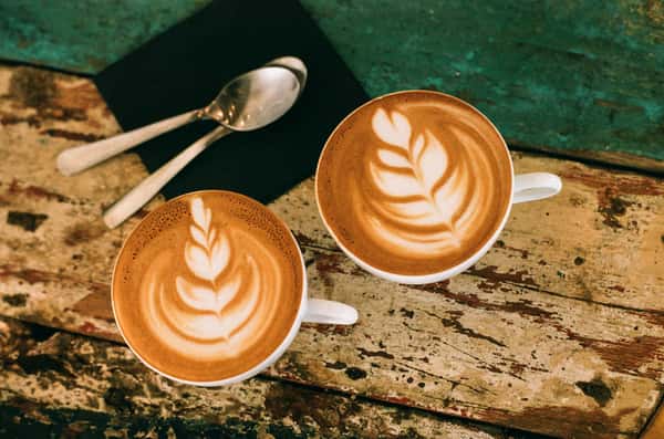 Two cups of latte with rosetta latte art on a rustic wooden table, accompanied by two spoons on a dark napkin.