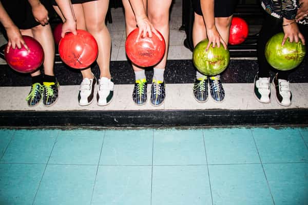 Five people in bowling shoes holding colorful bowling balls, standing at the edge of a lane with bright blue flooring.