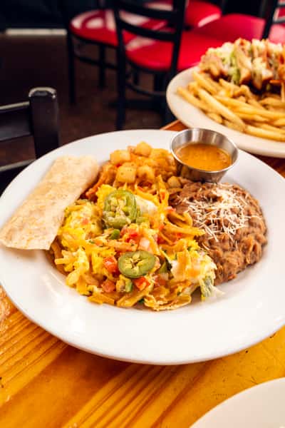 Vibrant Mexican breakfast plate featuring scrambled eggs with jalapeños, refried beans, potatoes, and a tortilla, served on a warm wooden table in a restaurant.