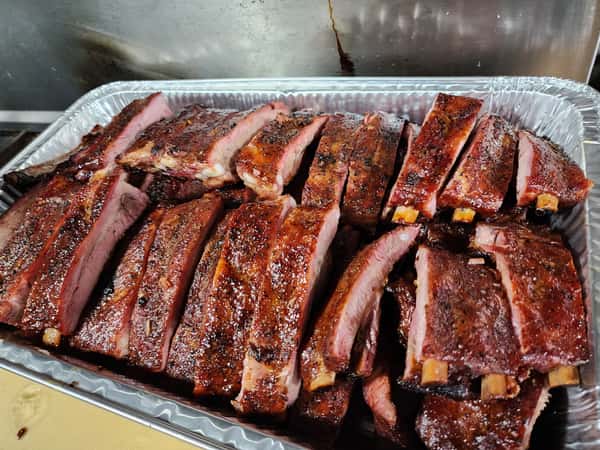 Tray of juicy, smoked pork ribs with a glossy glaze and charred edges, stacked in a disposable aluminum pan.