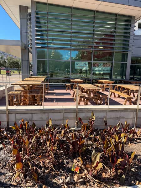Outdoor dining area with wooden picnic tables in front of a modern glass-front restaurant, with a "Now Hiring" sign in the window.