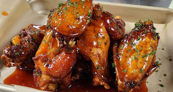 Sticky glazed chicken wings coated in a sweet and tangy barbecue sauce, garnished with chopped herbs in a takeout tray.