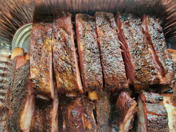 Tray of sliced, dry-rubbed pork ribs with a caramelized crust and smoky finish, arranged in a disposable aluminum pan.
