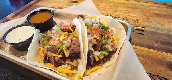 Two brisket tacos filled with chopped smoked beef, shredded cheese, diced onions, and jalapeños, served with BBQ and ranch dipping sauces.
