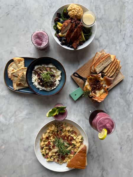 overhead restaurant spread with grilled chicken salad, creamy pasta, baked brie with bread, hummus and pita, and fresh berry cocktails on a marble table