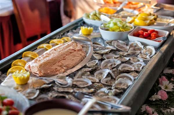 Chilled oysters on the half shell on top of bed of ice beside a plate of salmon decorated with lemon wedges next to bowls of assorted fruits and vegetables