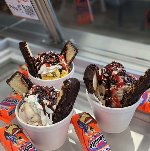 Three cups filled with ice cream topped with Gansito cake bars, whipped cream, chocolate syrup, sprinkles, and fruit toppings.