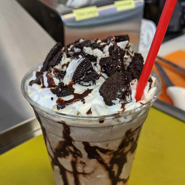 A chocolate milkshake topped with whipped cream, Oreo cookie pieces, and chocolate syrup, served with a red straw.