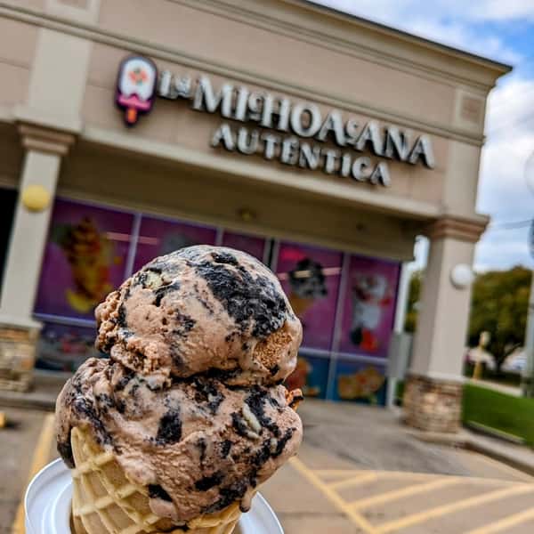 A waffle cone with two scoops of cookies and cream ice cream, with the "La Michoacana Autentica" shop in the background.