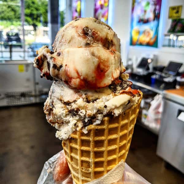 A waffle cone filled with two scoops of ice cream, one with chocolate and the other with strawberry, drizzled with syrup.