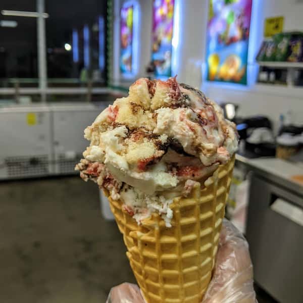 A cone of ice cream topped with crumbled cake pieces and swirled with fruit and chocolate syrup.