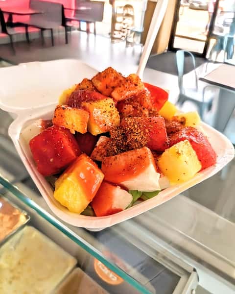 A container filled with colorful fresh fruit, including watermelon and pineapple, topped with chili powder and seasoning.