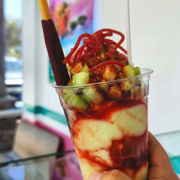 A colorful fruit and snack cup filled with mango, cucumber, and other fruits, topped with chili powder, a tamarind candy stick, and sweet syrup.